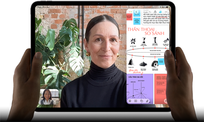 iPad Pro, vỏ ngoài mặt trước, nằm ngang, màu đen không gian, các góc bo tròn, viền màn hình màu đen, bàn tay cầm ở bên trái và bên phải, màn hình hiển thị cuộc trò chuyện FaceTime đang được dịch trực tiếp, và một ứng dụng giáo dục hiển thị bài thuyết trình "Thần Thoại So Sánh" với văn bản, biểu đồ và ghi chú viết tay